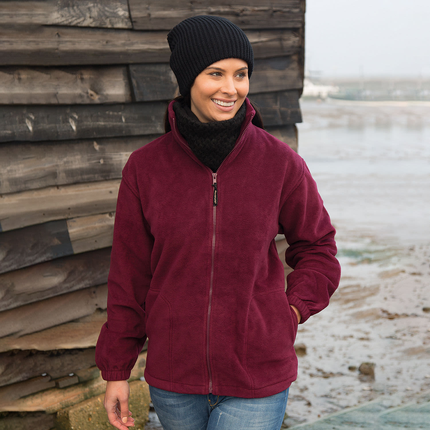 A maroon polar fleece jumper with a full zip, worn by a woman in a black beanie against a rustic backdrop.
