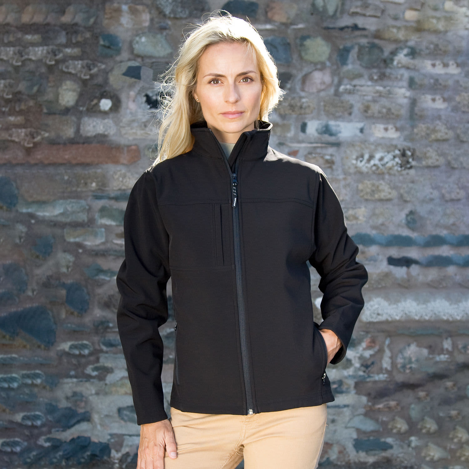 A black ladies' classic softshell jacket with a zip front and a chest pocket, worn by a model against a stone wall.