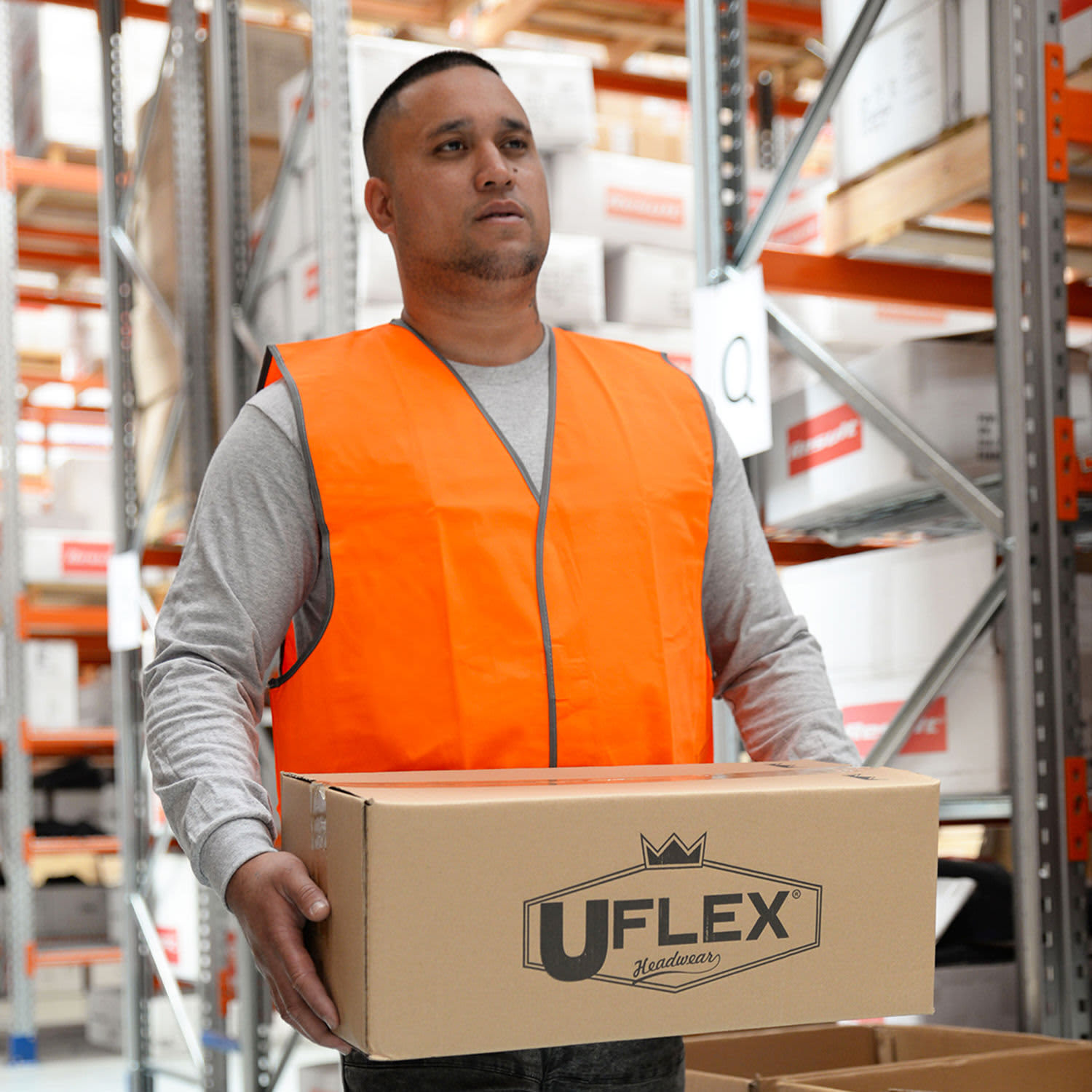 A man wearing an orange high-visibility safety vest holds a cardboard box in a warehouse setting.