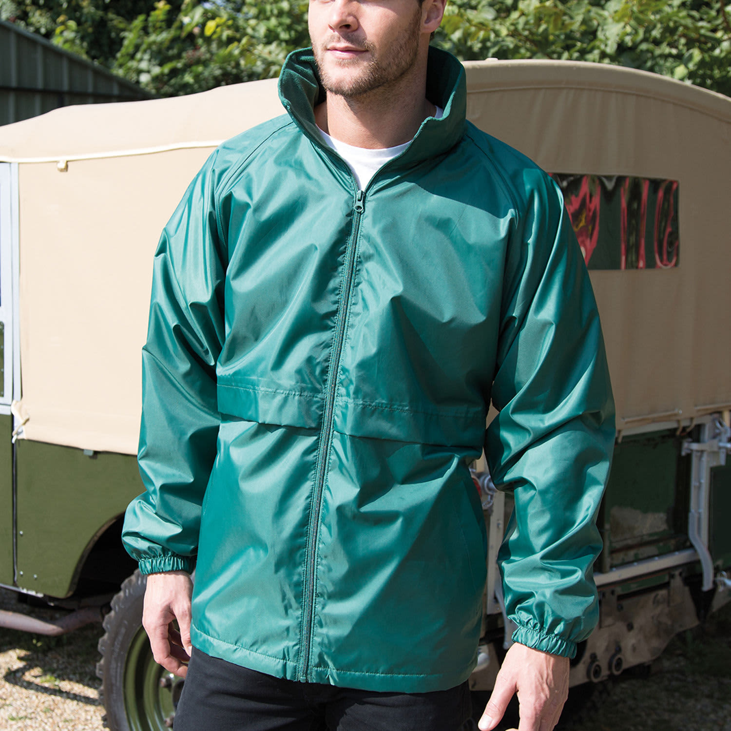 A green rain jacket featuring a high collar and elastic cuffs, modelled by a man outdoors. It has a logo on the chest.