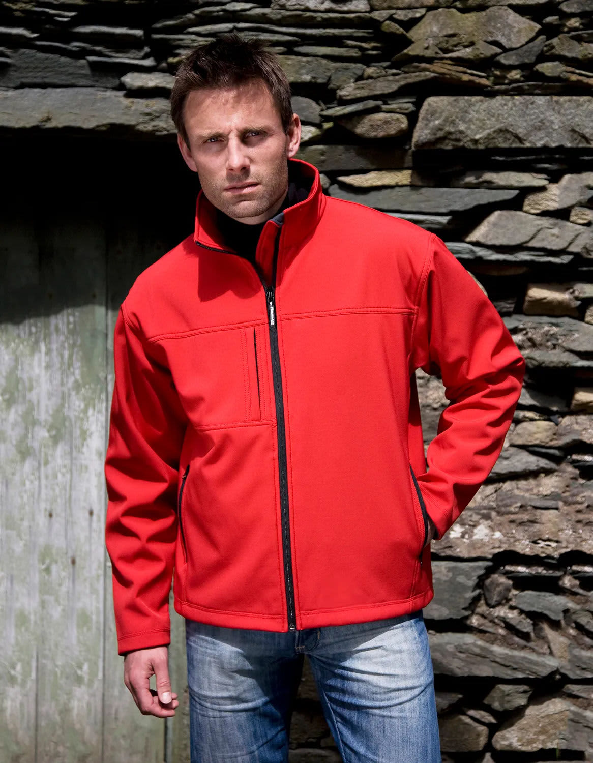 A red soft shell jacket is worn by a model standing against a stone wall, featuring a zip and a slight collar.
