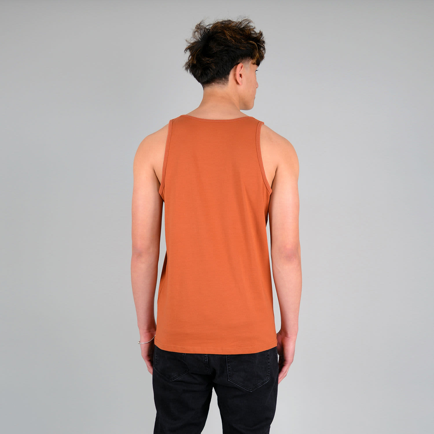 An adult male model wearing a brown singlet from the back, with dark pants and a plain background.