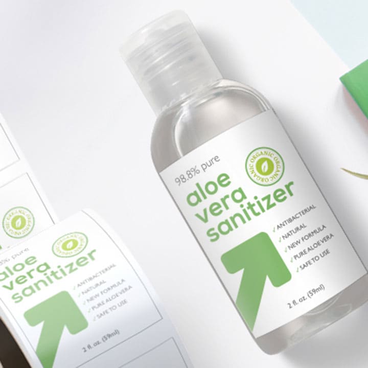 A clear bottle of aloe vera sanitiser with a green label and branding, surrounded by printed sticker rolls.