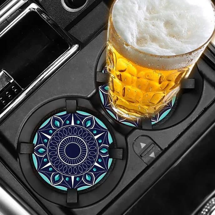 A drink coaster featuring a vibrant blue and turquoise mandala design, placed in a car cup holder alongside a glass of beer.
