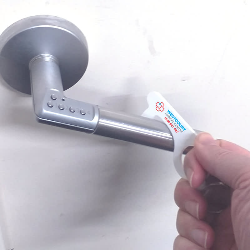 A hand holding a white Covid Key tool designed for touching surfaces, shown on a silver door handle.