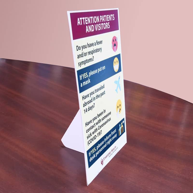A freestanding sign in white and purple on a wooden table, displaying health check questions for patients and visitors.