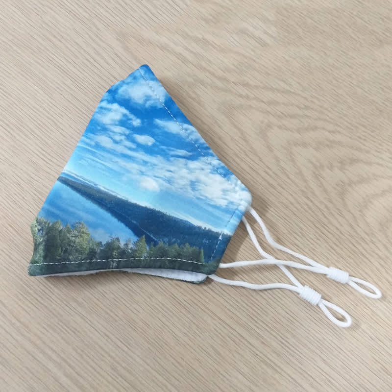 A fabric face mask featuring a scenic blue sky and landscape design with adjustable white ear straps.