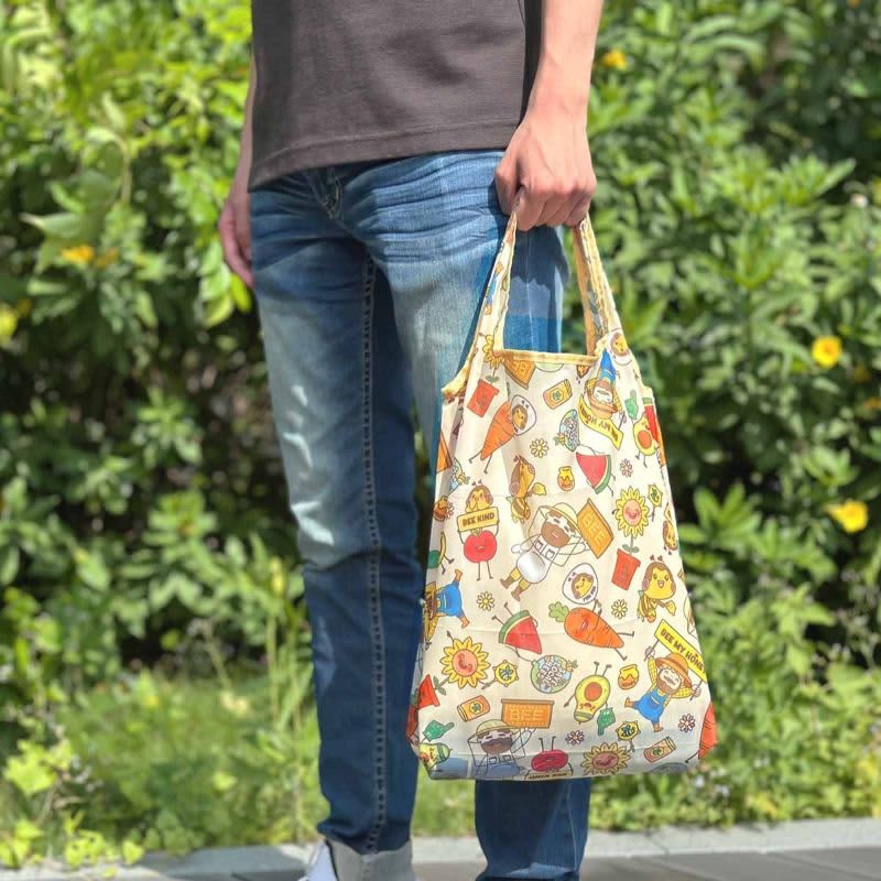 A foldable shopping bag in cream with colourful food-themed designs, held by a person in casual clothing.