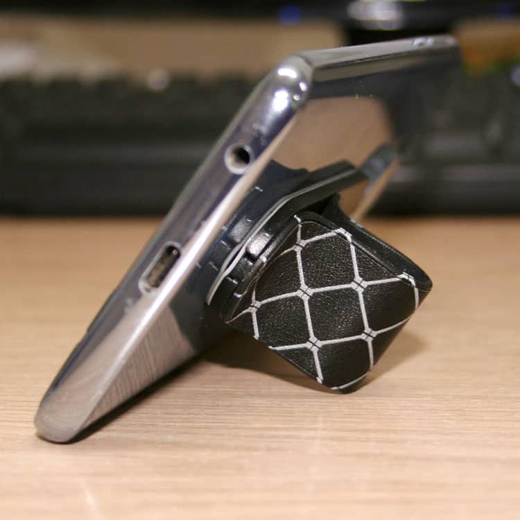 A phone stand in black with a geometric pattern, supporting a smartphone at an angle on a wooden surface.