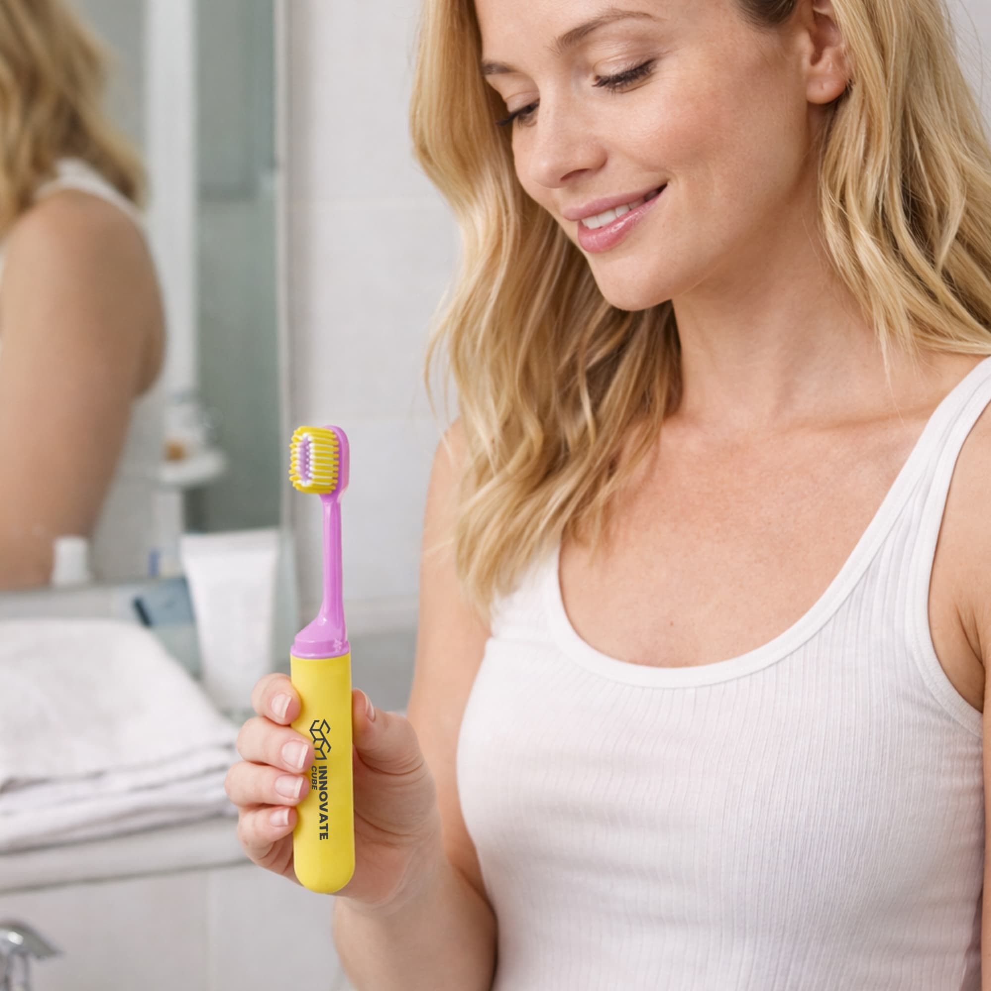 A pink and yellow foldable soft bristle toothbrush held by a woman in a bathroom setting, with a mirror in the background.