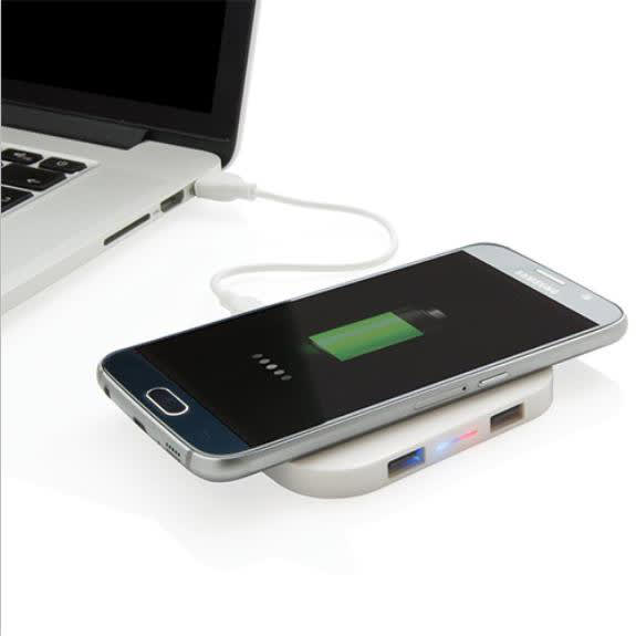 A square-shaped wireless charger in white, with a smartphone displaying a charging battery, next to a laptop.