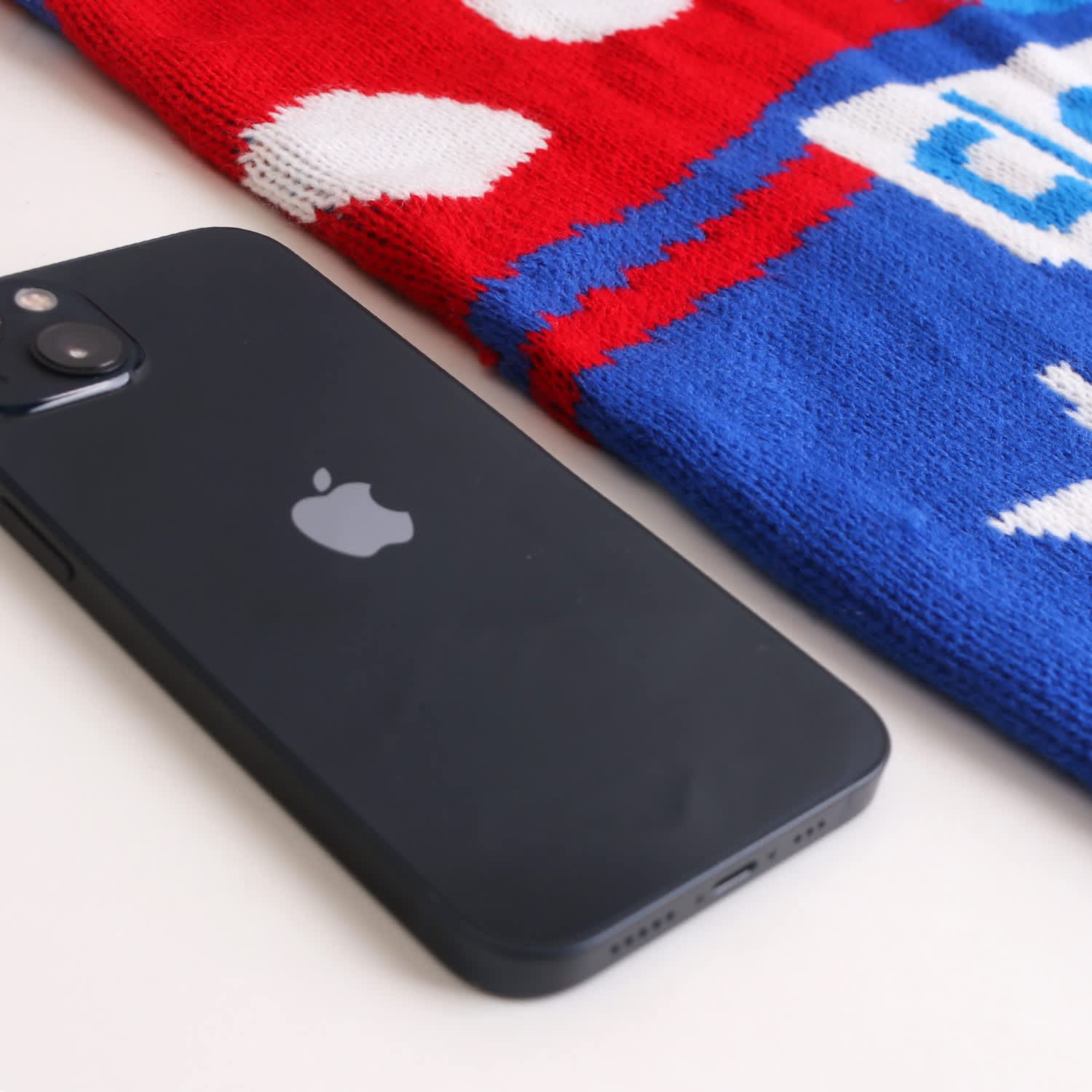 A black smartphone lies beside a red and blue jacquard sweater, featuring a playful pattern. The sweater has a logo.