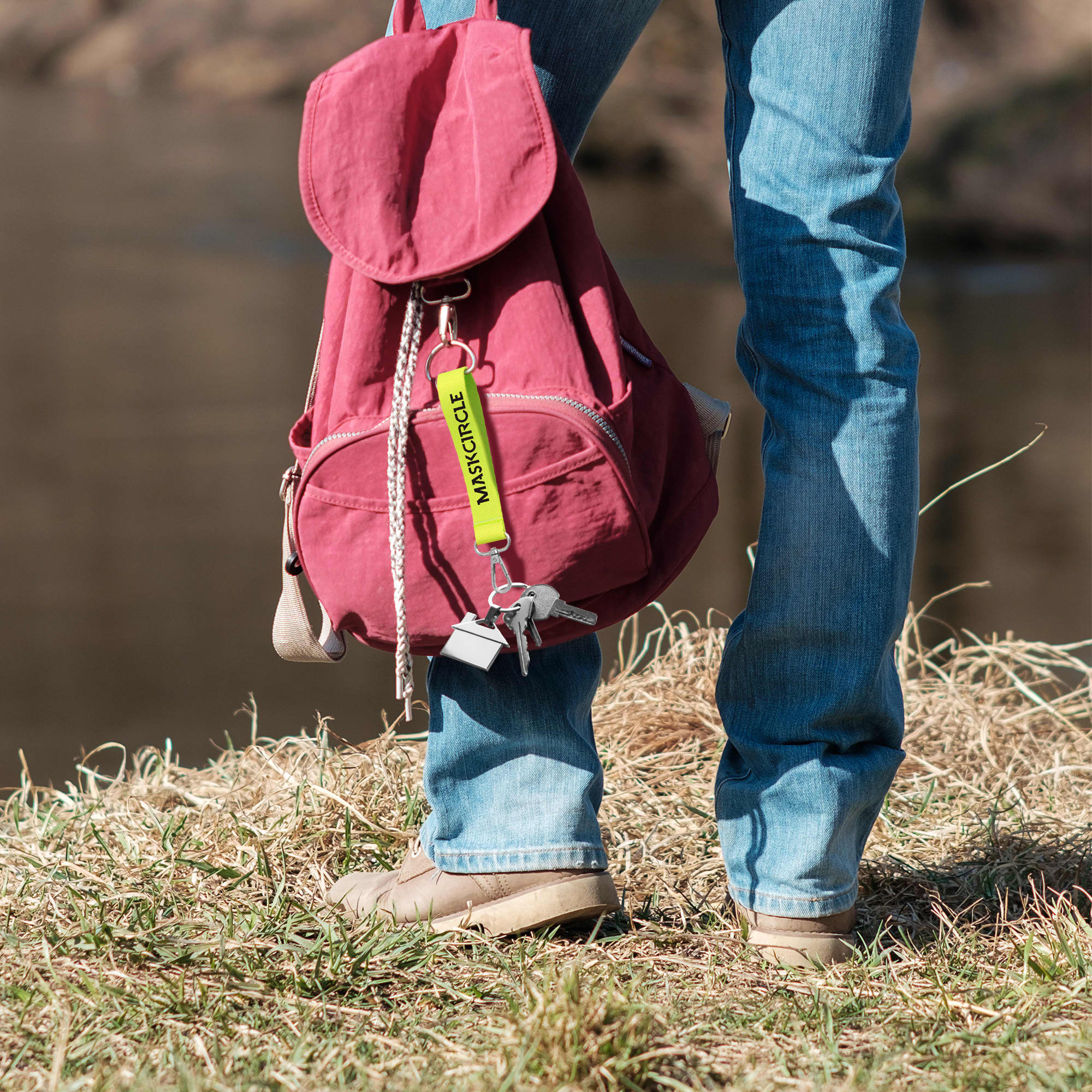 A keyring in yellow attached to a maroon backpack, held by a person standing on grass near a riverbank.