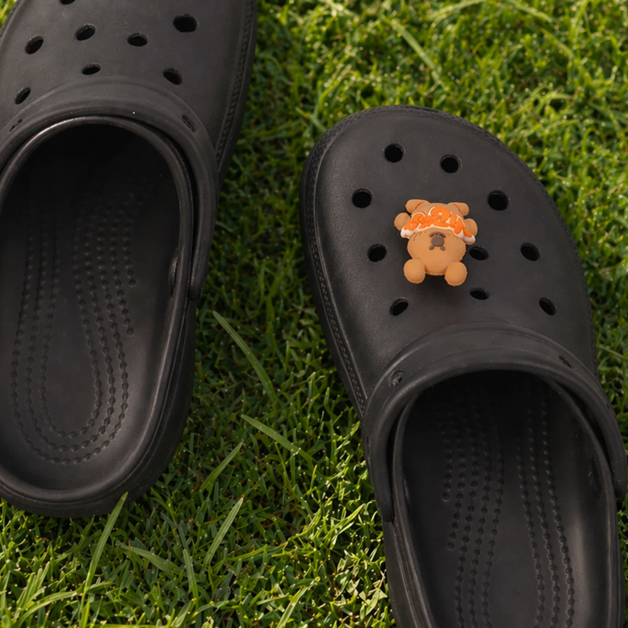 A pair of black clog shoes featuring a 3D bear charm on one shoe, set on green grass.