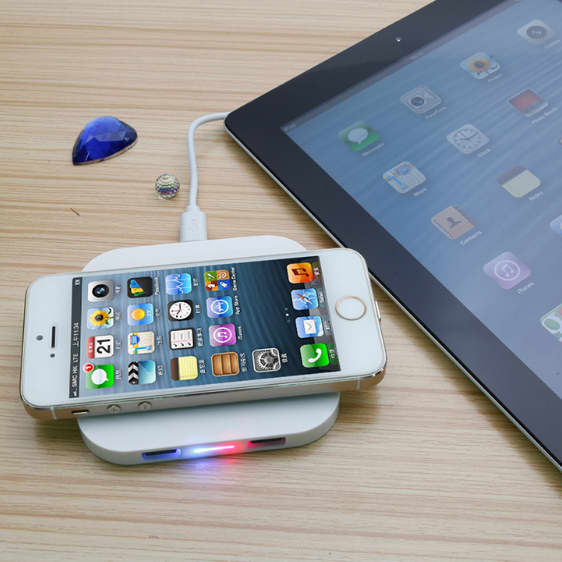 A square-shaped wireless charger in white, with a smartphone placed on top, beside a tablet on a wooden surface.