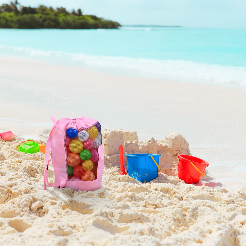 A pink foldable mesh drawstring backpack filled with colourful balls sits on the beach beside buckets and a sandcastle.