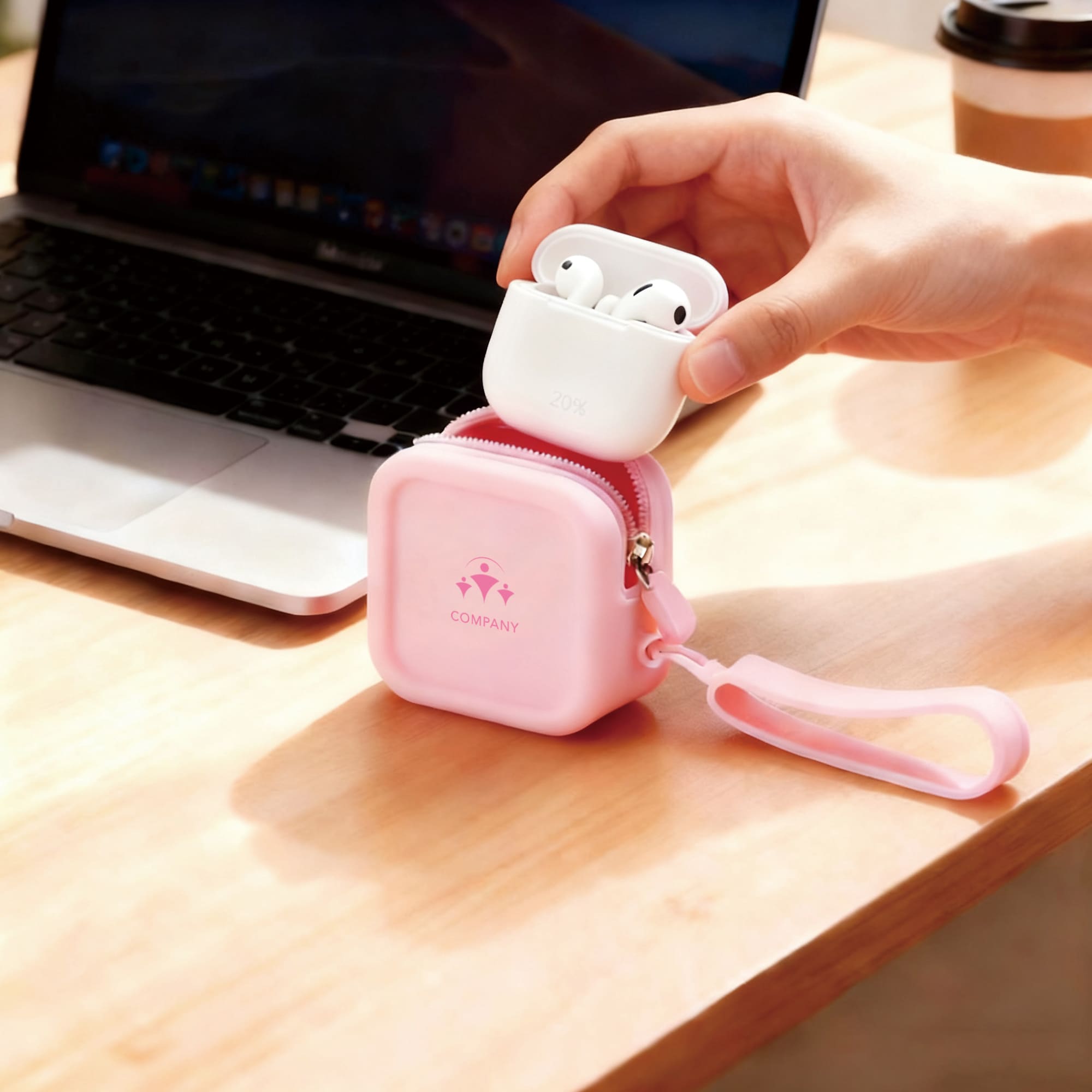A pink square silicone coin pouch with a zip, holding a white wireless earbud case, next to a laptop.