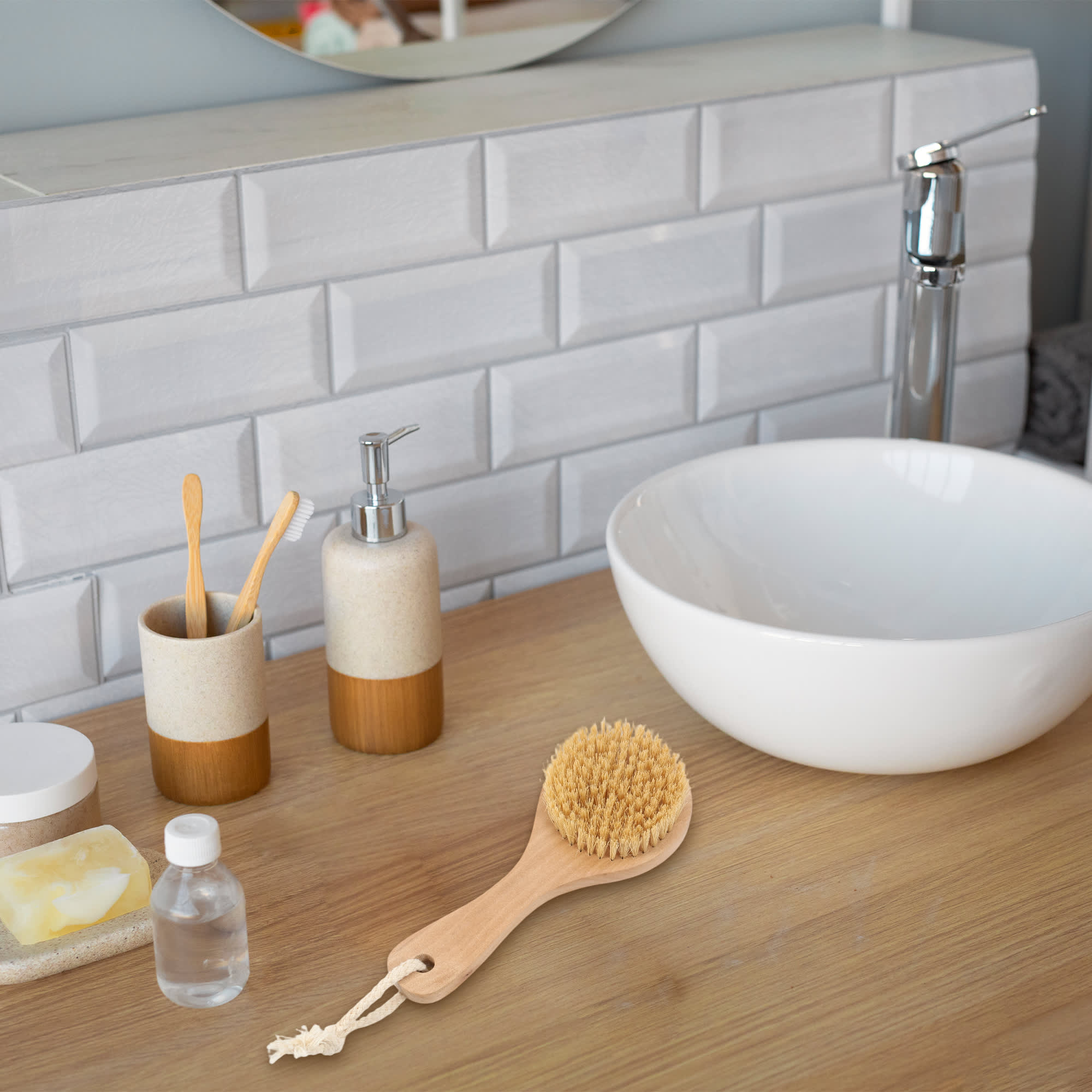 A round body brush with a short wooden handle sits on a wooden countertop near a white bowl and various bathroom items.