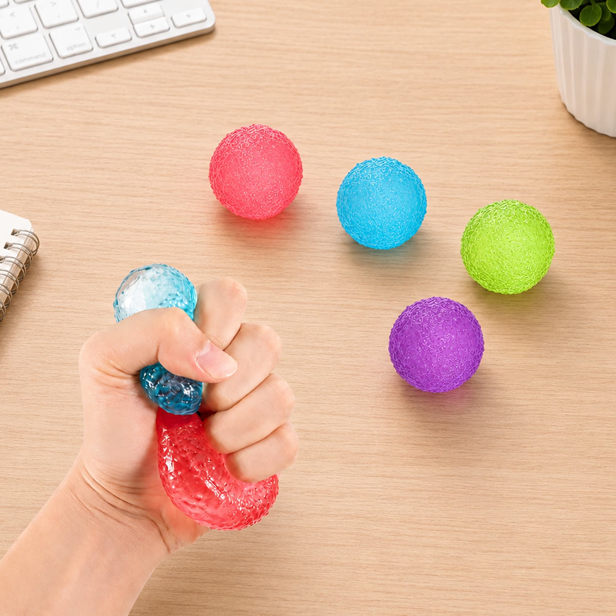A hand squeezing a clear squishy stress relief mini ball, surrounded by coloured stress balls in red, blue, green, and purple.