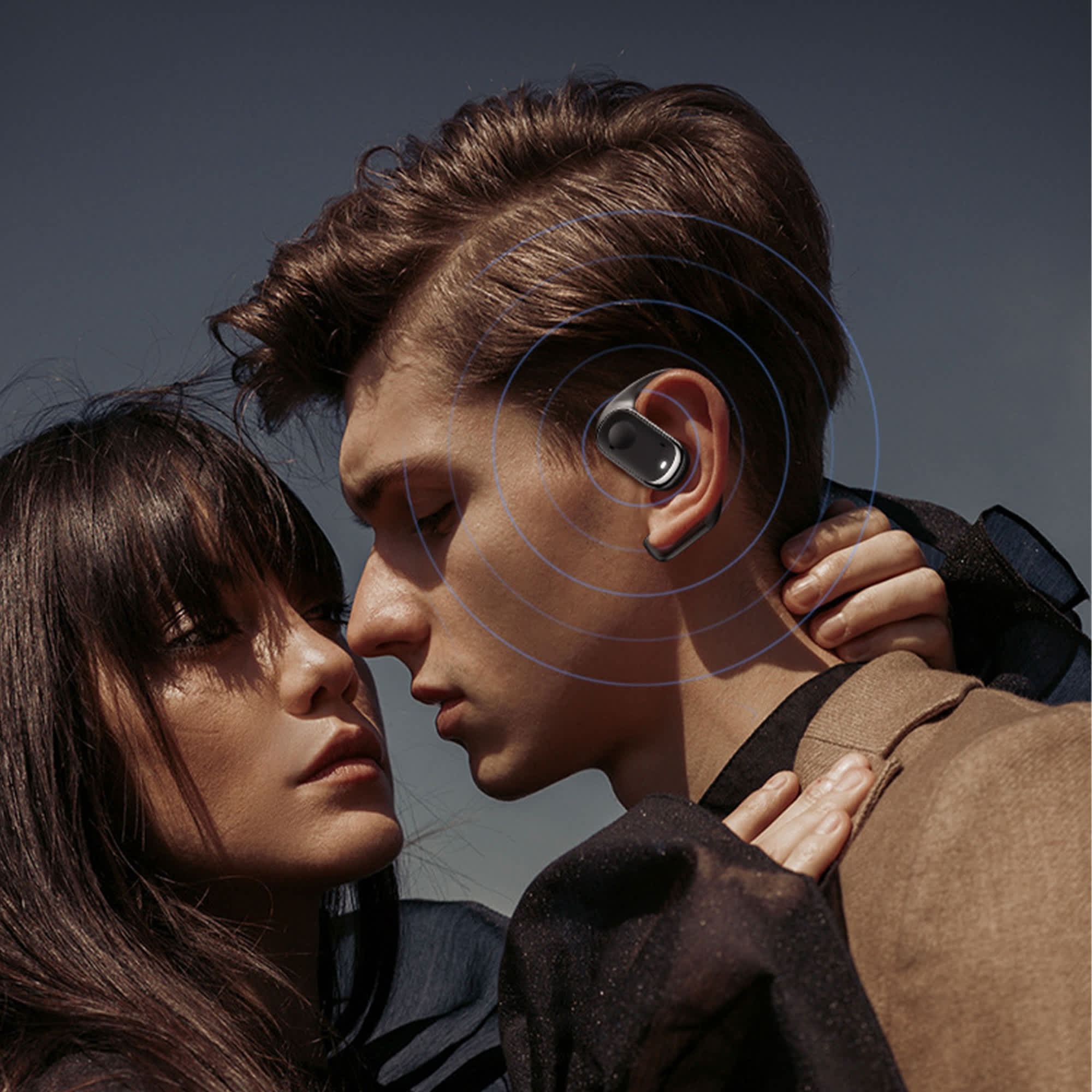 Open-ear headphones in silver are worn by a person, who is facing another person, against a clear sky backdrop.