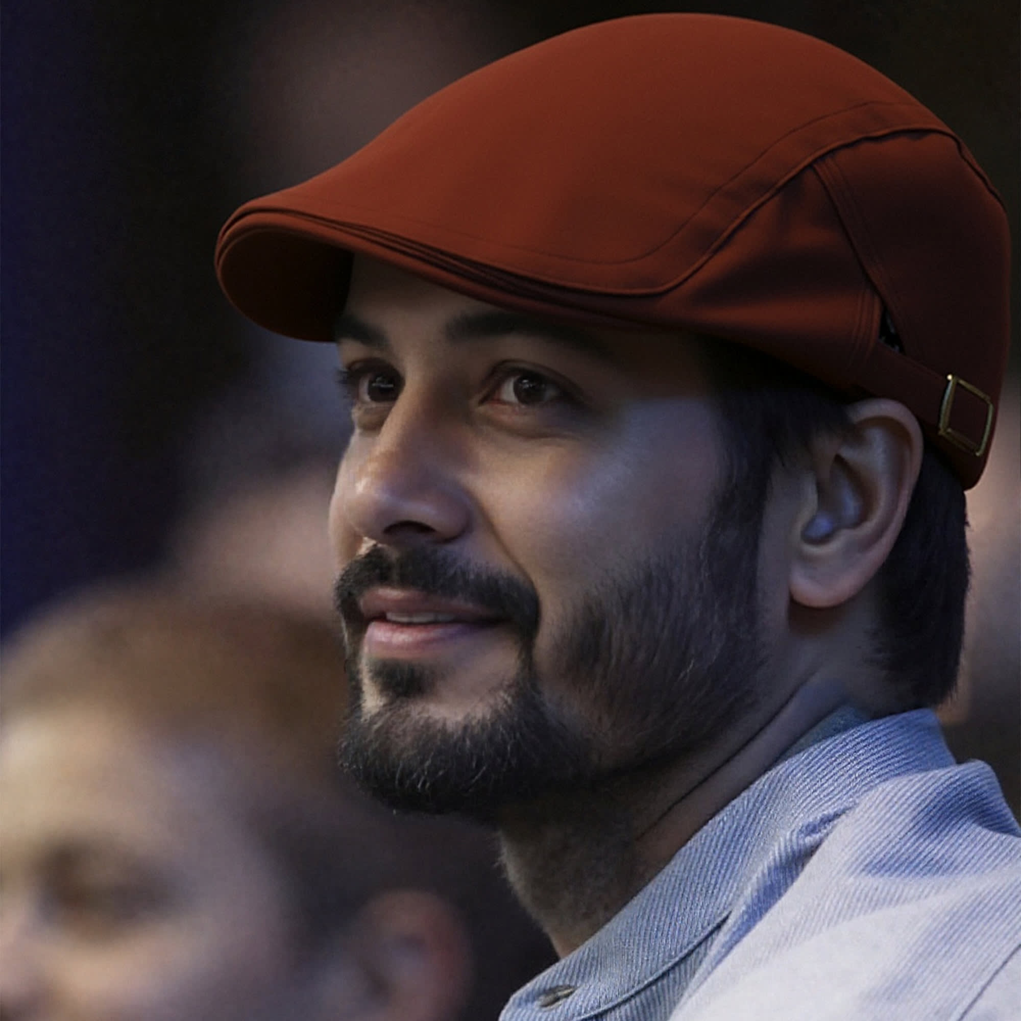 A man wearing a burgundy beret hat smiles, with a blurred crowd in the background. The hat has a logo.