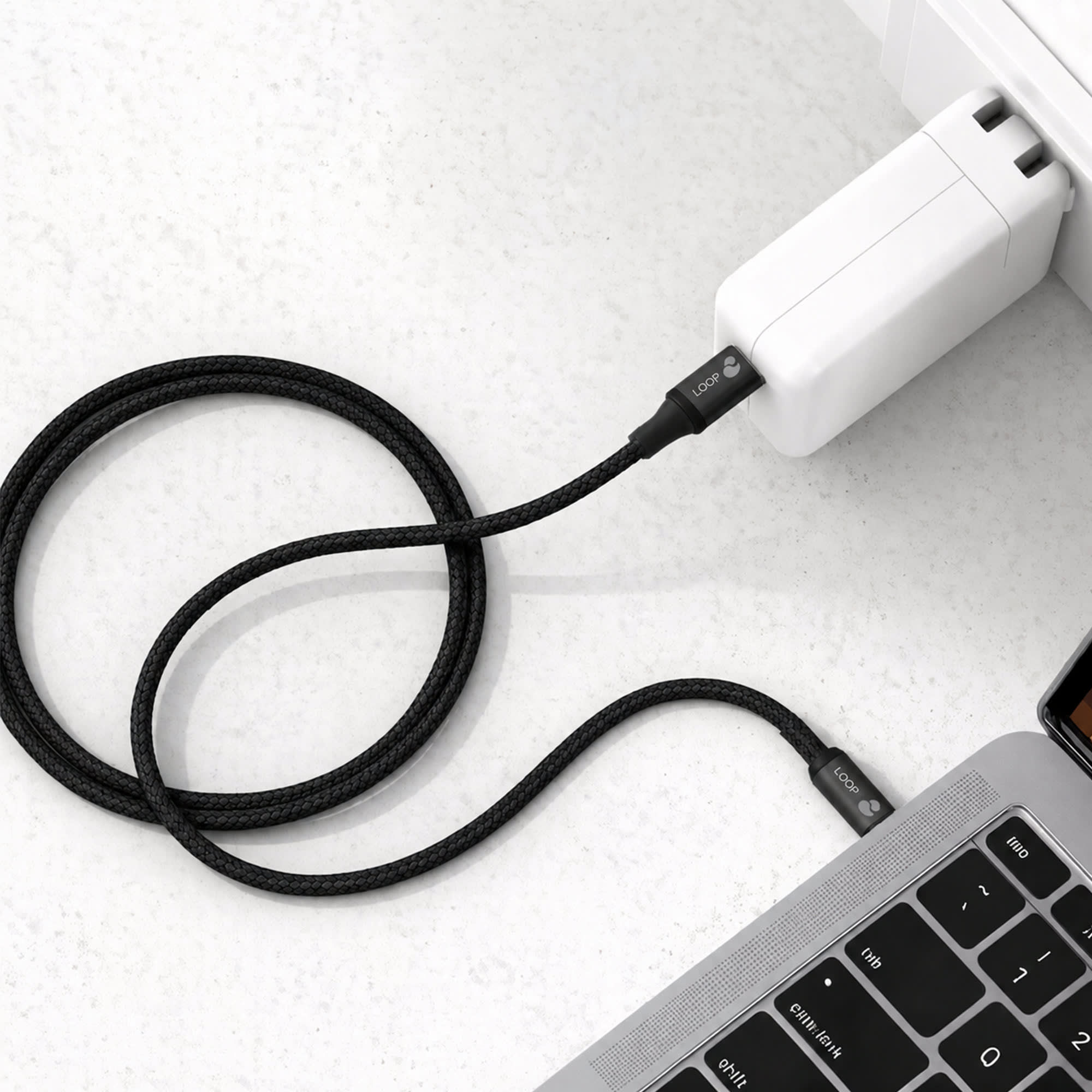 A black braided charging cable connected to a white power adapter and a laptop. It features a logo on the cable.