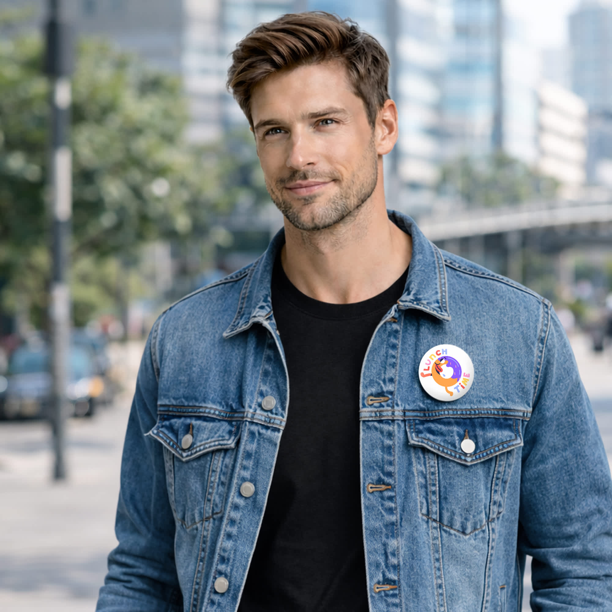 A man wearing a denim jacket and a button badge stands outdoors in an urban setting, with greenery and buildings in the background.