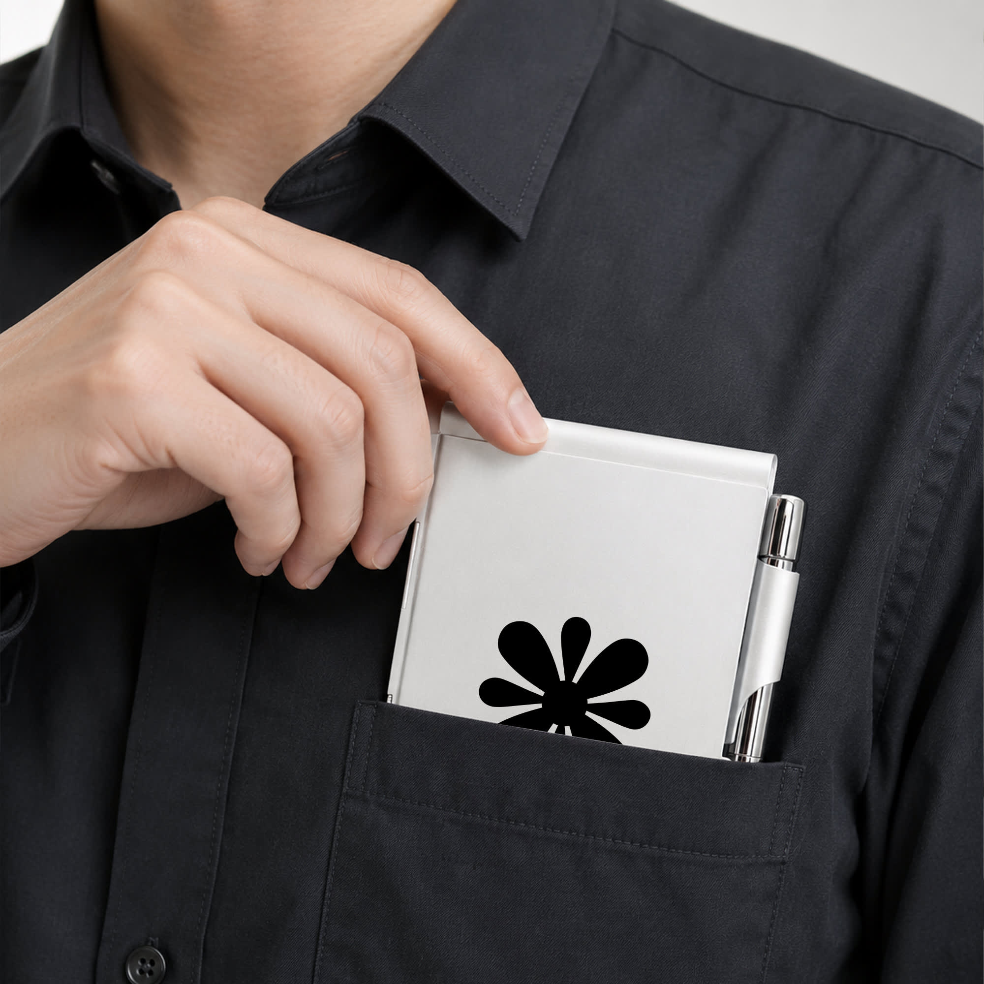 A white aluminium pocket notebook being placed into a shirt pocket, alongside a pen. The notebook features a logo design.