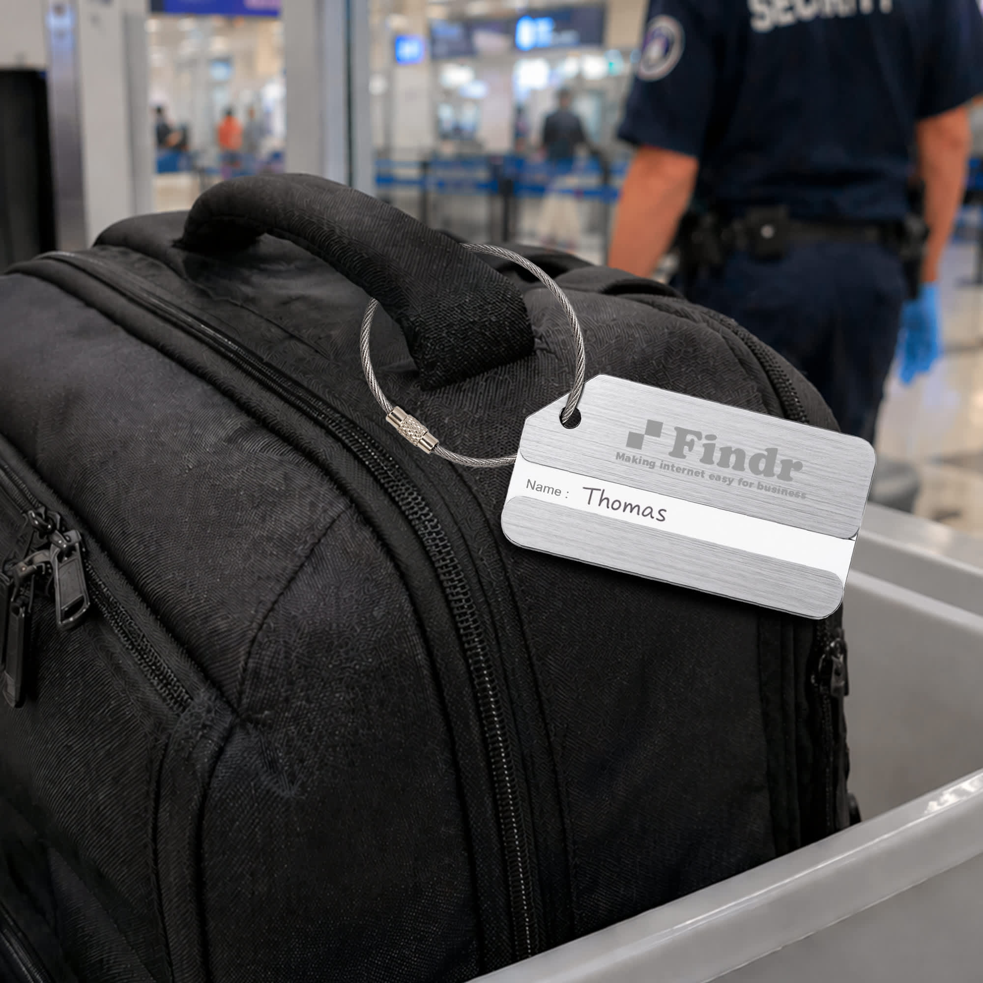 A square brushed aluminium luggage tag attached to a black bag, featuring a logo and a name label.
