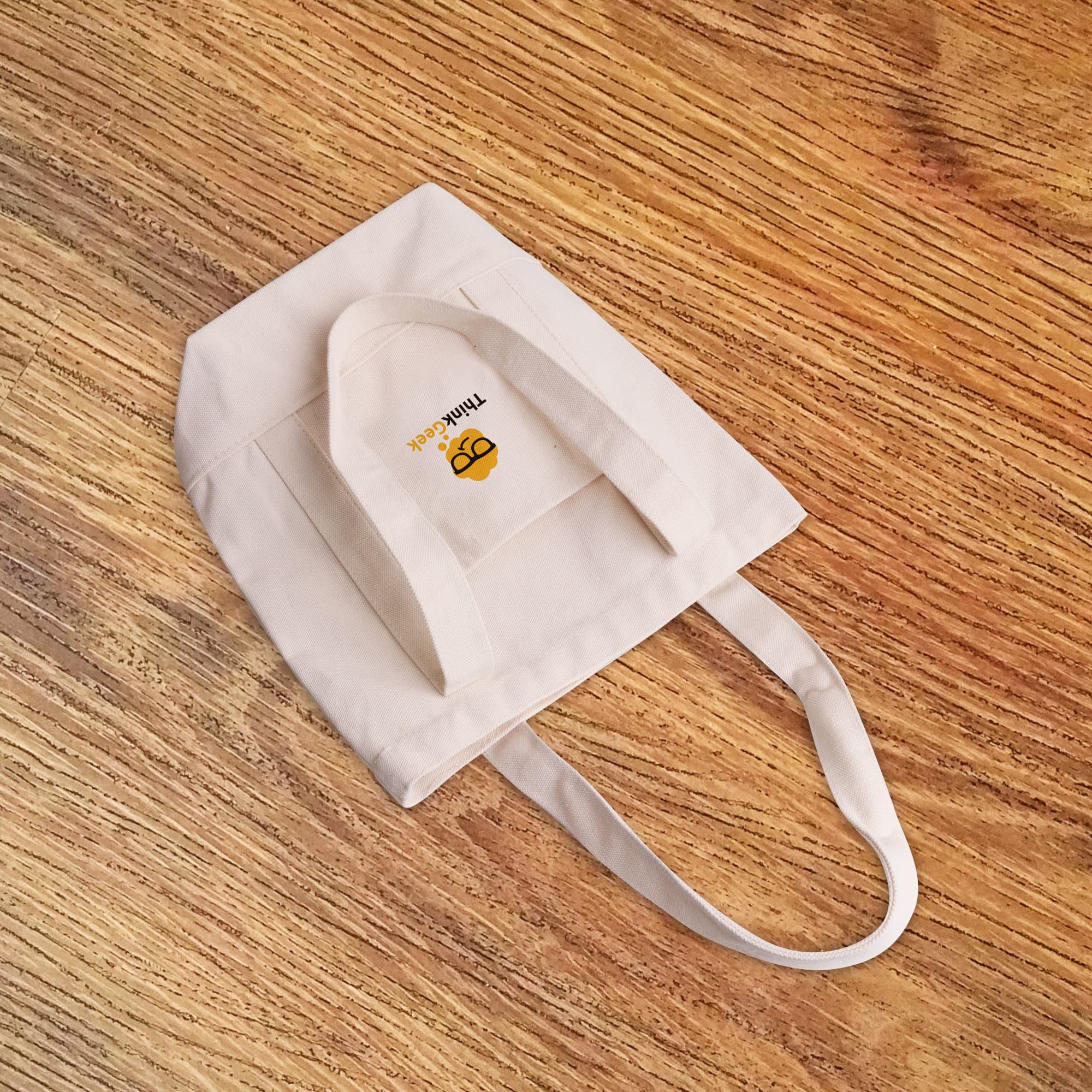 A natural-coloured, heavy-duty cotton tote bag with handles and a logo. The bag is placed on a wooden surface.