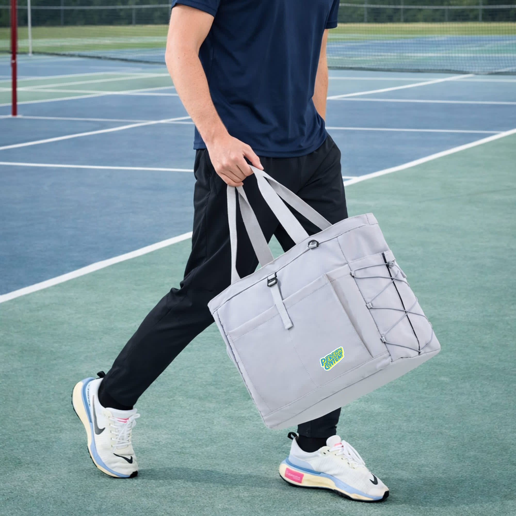 A grey lightweight tennis tote bag with handles, featuring a logo and elastic bungee cord, carried by a person on a tennis court.