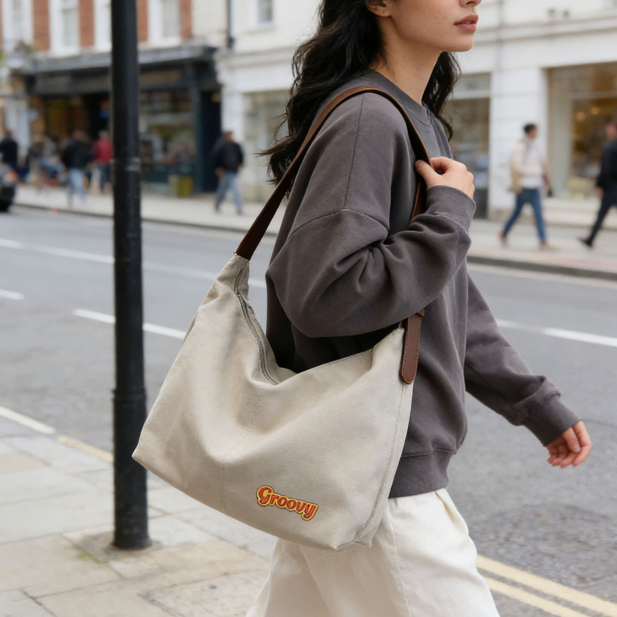 A beige canvas tote bag with brown straps, carried by a person in a grey sweater while walking on a city street.