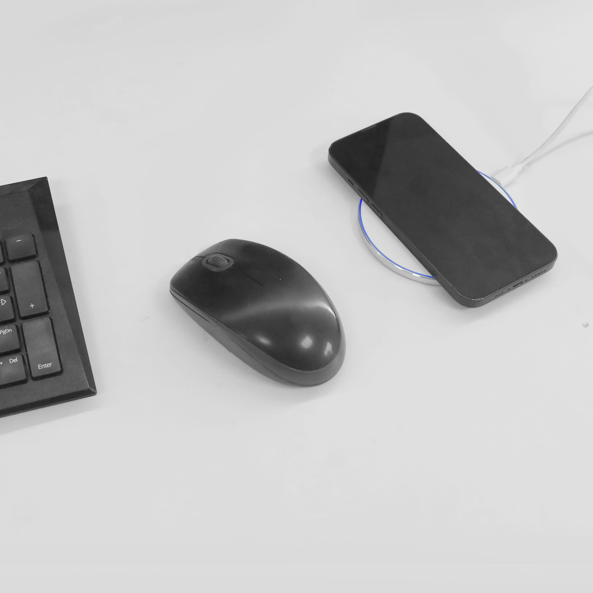 A round black wireless charger with a smartphone on it, positioned next to a black computer mouse and keyboard.