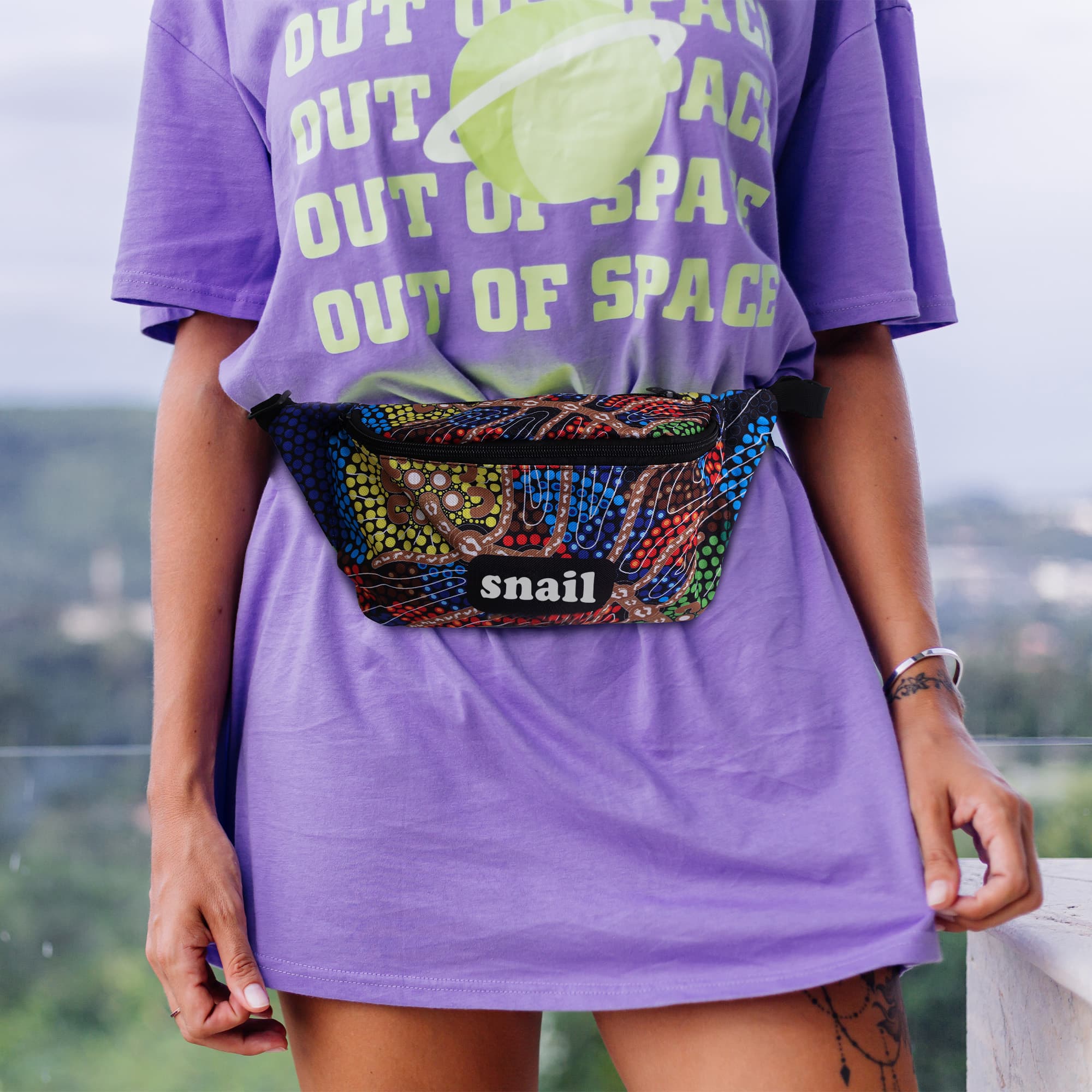 A colourful sublimation bum bag featuring a mix of vibrant patterns, worn around the waist of a person in a purple shirt.