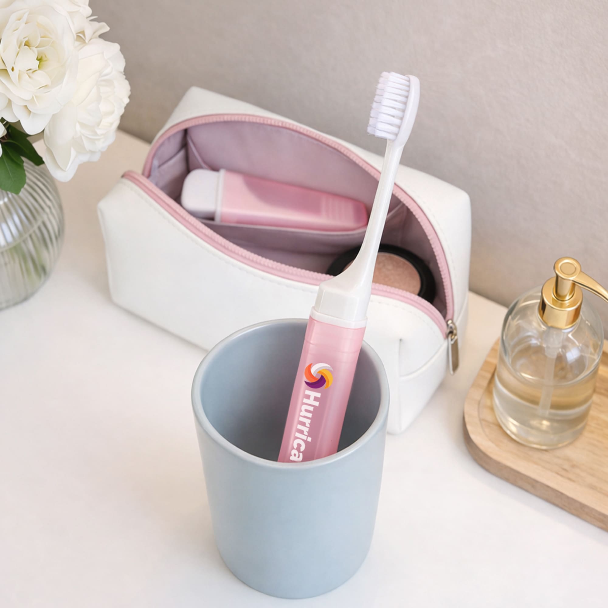 A pink transparent foldable handle toothbrush is placed in a grey cup next to a white and pink cosmetic bag and a glass bottle.