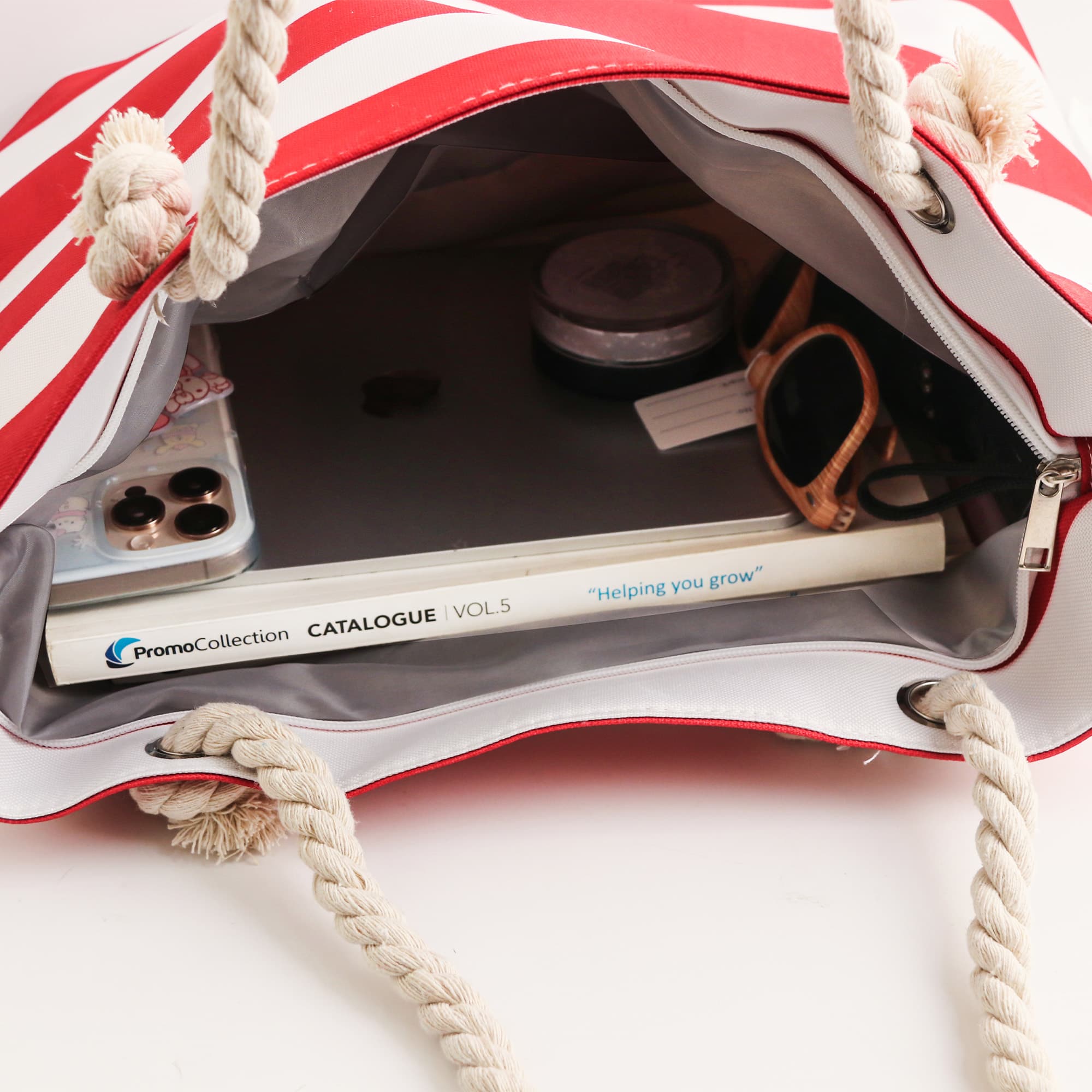 A tote bag with red and white stripes featuring a rope handle, containing a phone, sunglasses, and a catalogue.