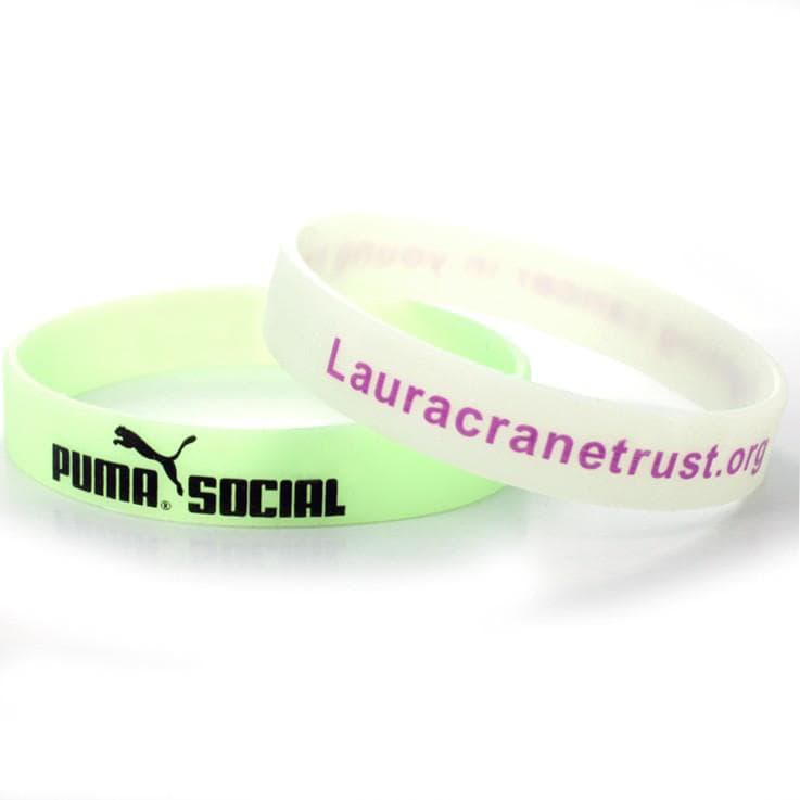 Two glow-in-the-dark silicone wristbands, one in pale green and the other white, featuring a logo on the green band.