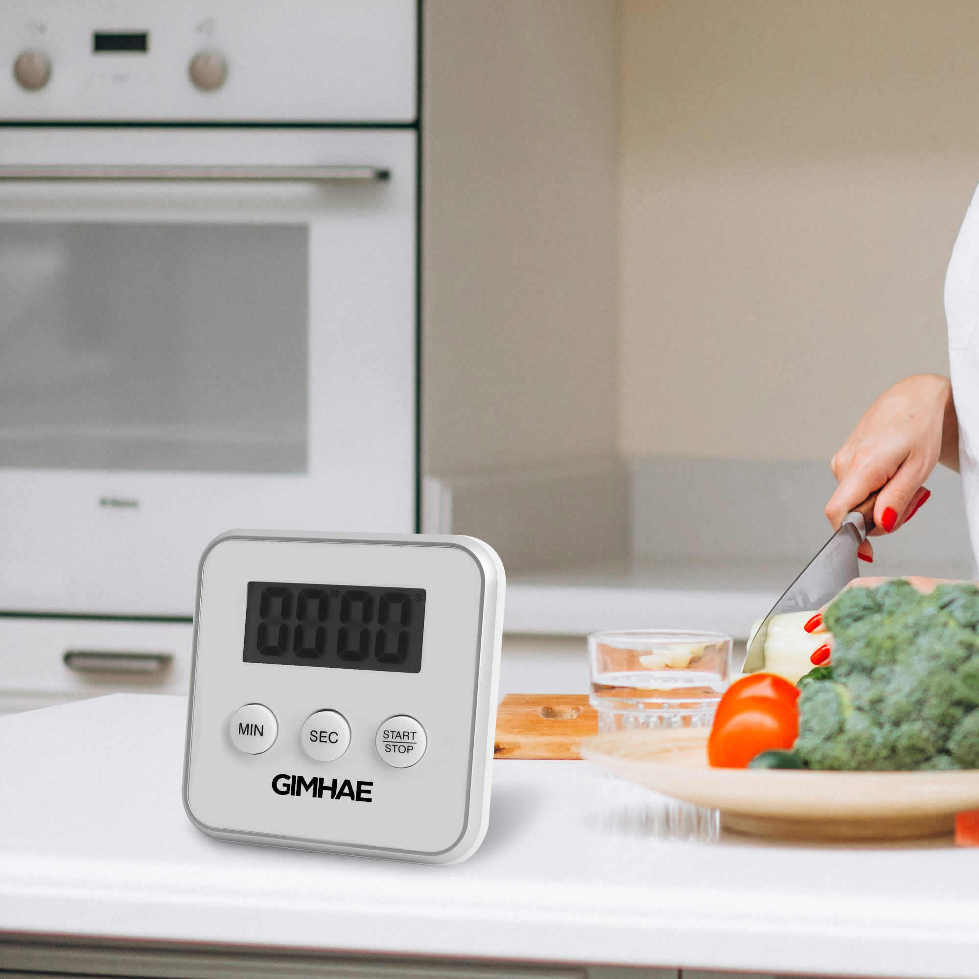 A digital kitchen timer in white with a large display, placed on a kitchen countertop beside fresh vegetables.