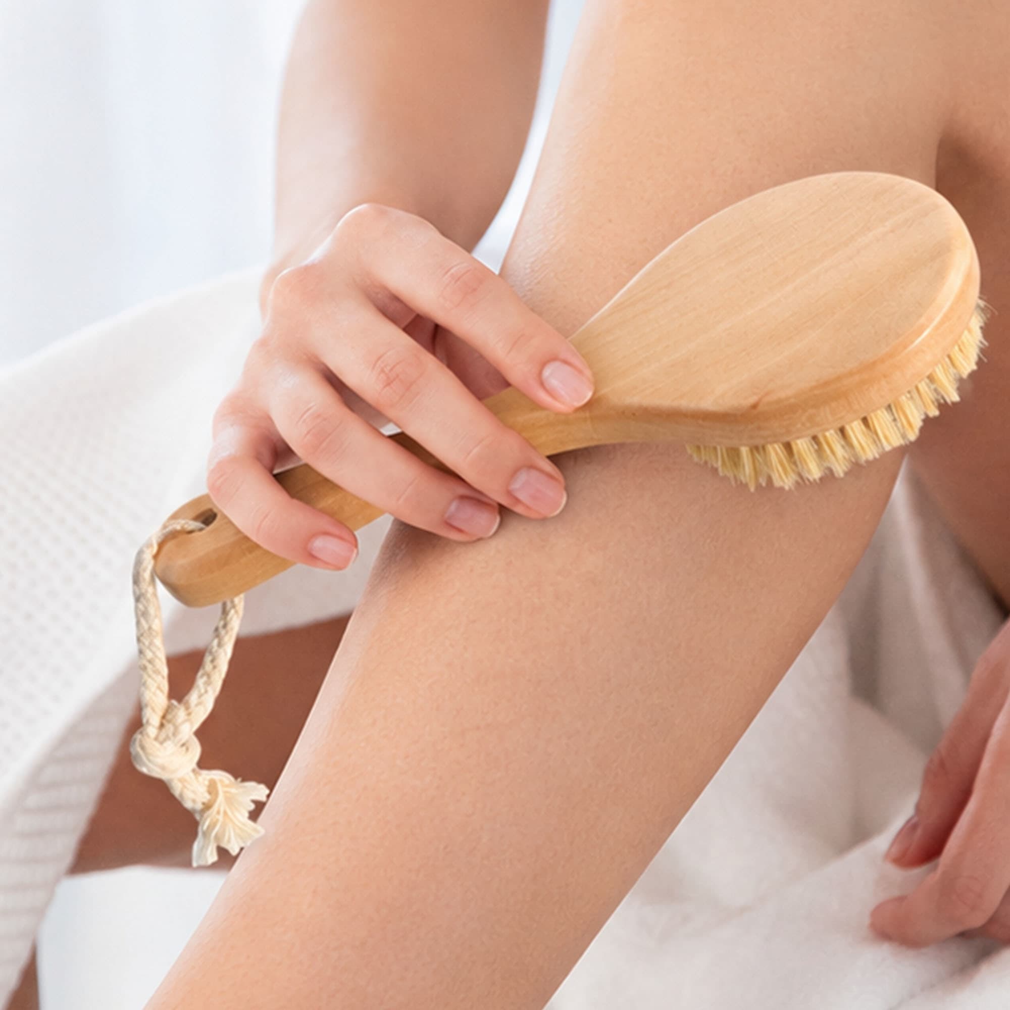 A round body brush with a short wooden handle and natural bristles, being used on skin, with a rope loop for hanging.