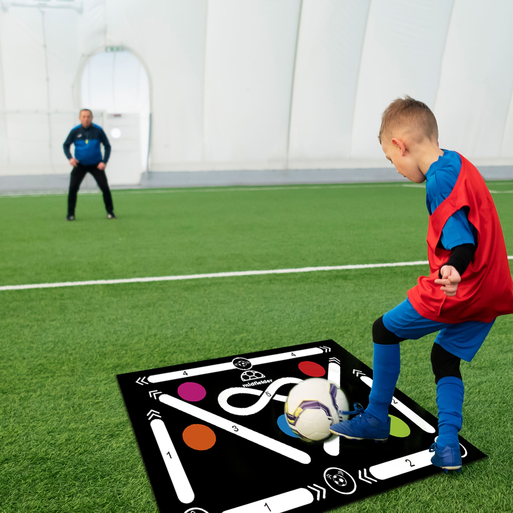 A black football training mat with white lines and colourful circles, placed on green turf, with a player and coach visible.