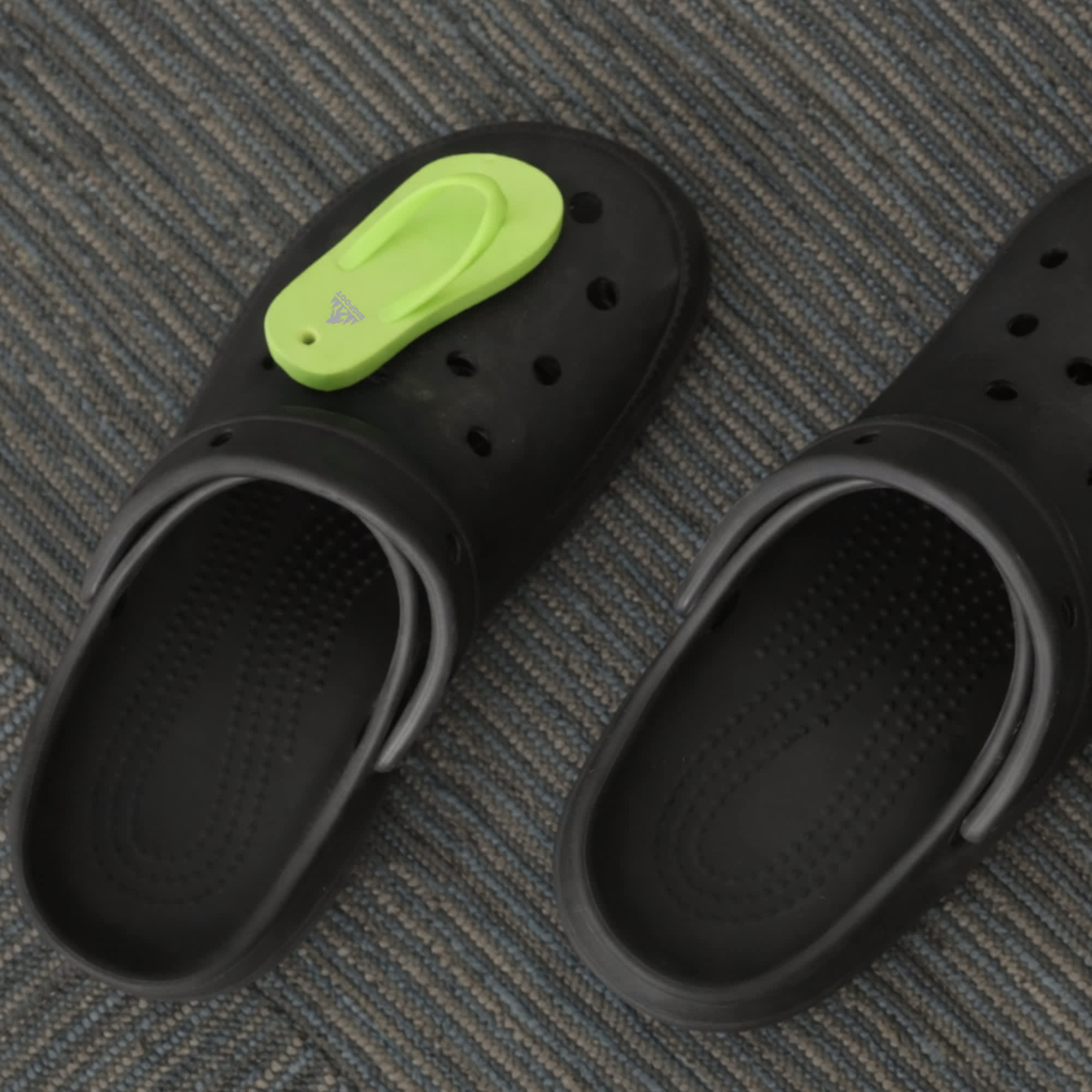 A pair of black clog shoes with a bright green thongs-shaped charm attached to one.