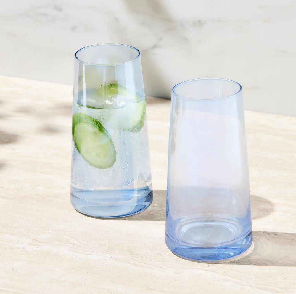 Two highball glasses in light blue, one filled with water and cucumber slices, displayed on a marble surface.