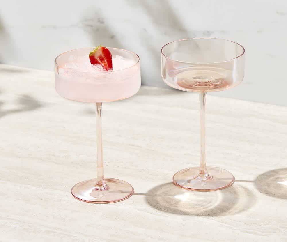 A set of two pink coupe glasses, one filled with a drink and garnished with a strawberry, displayed on a light surface.