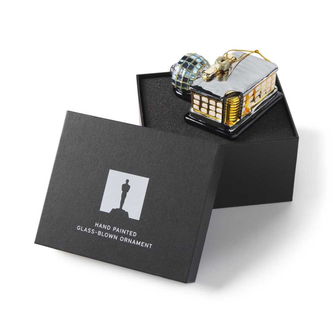 A detailed glass-blown ornament in a black gift box, featuring a cabin design with golden accents and a glossy finish.