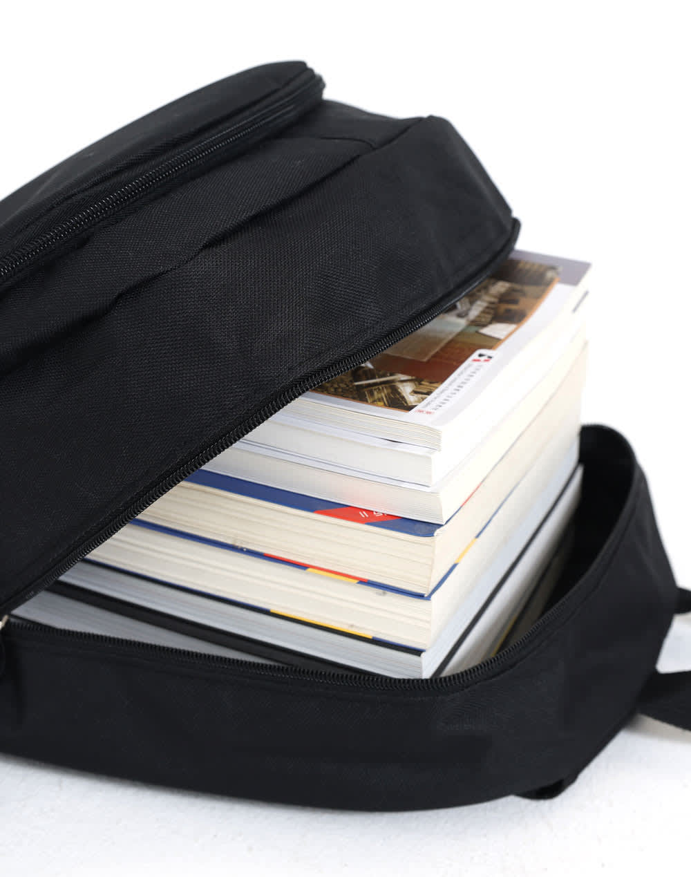 A black backpack partially open, revealing several stacked books inside. The backpack features a zip closure and a simple design.