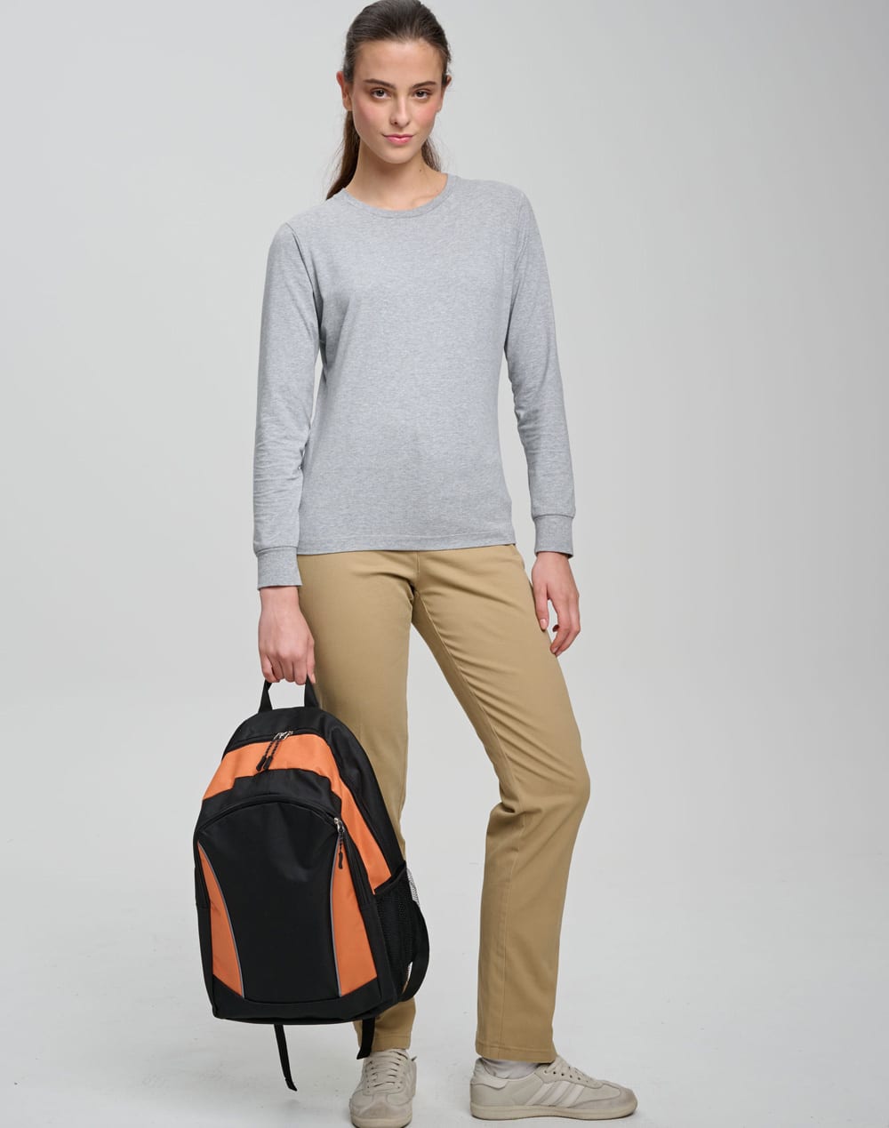 A young woman stands with an orange and black backpack, wearing a grey long-sleeve top and beige trousers.