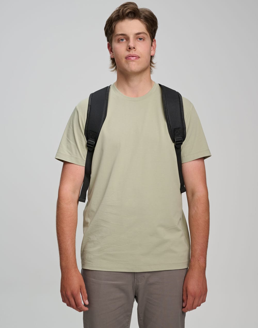 A man wearing a light khaki T-shirt and grey pants, carrying a black backpack with shoulder straps.