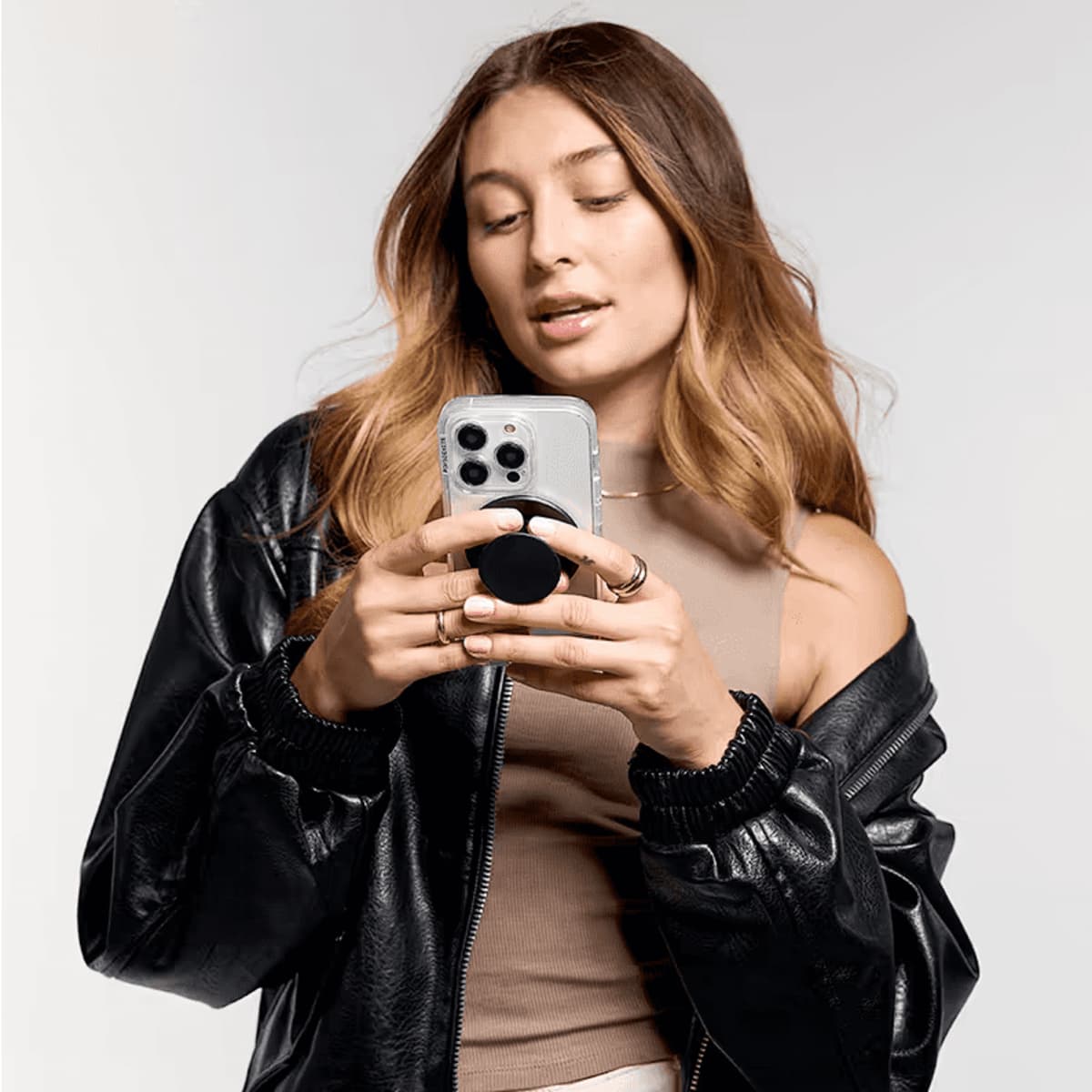 A person holding a smartphone with a PopSocket MagSafe attached, wearing a black leather jacket.