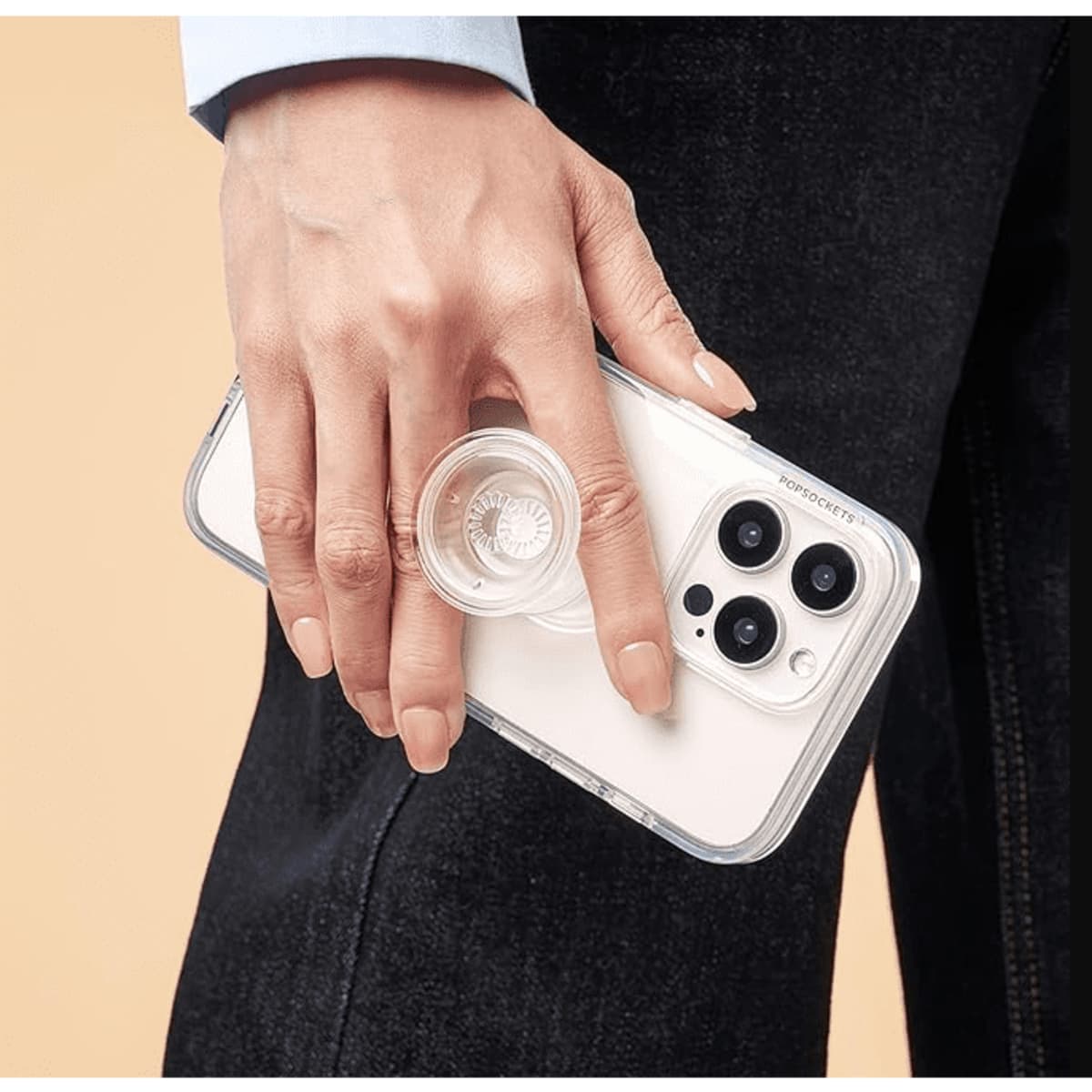 A hand holds a smartphone with a clear, circular PopSocket attached. The phone has a light-coloured case.