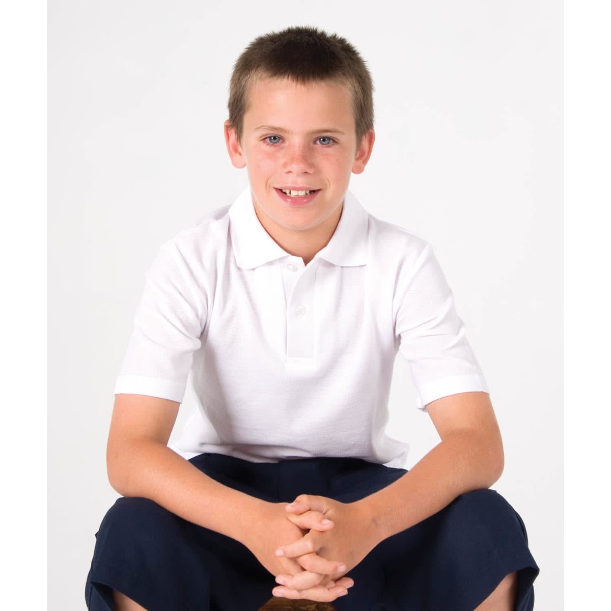 A white kid's pique polo shirt worn by a boy sitting, with dark shorts and a neutral background. The shirt has a logo.